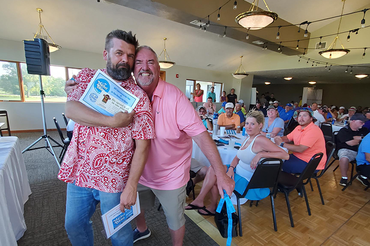 Two men standing together holding certificates, smiling at a gathering with many seated guests in a decorated banquet hall.