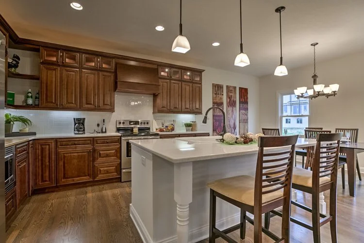 Kitchen with wooden cabinets, white subway tile backsplash, and stainless steel appliances. An island with a white countertop and three wooden chairs is in the center. Pendant lights hang above the island, and a dining table with chairs is nearby. Th