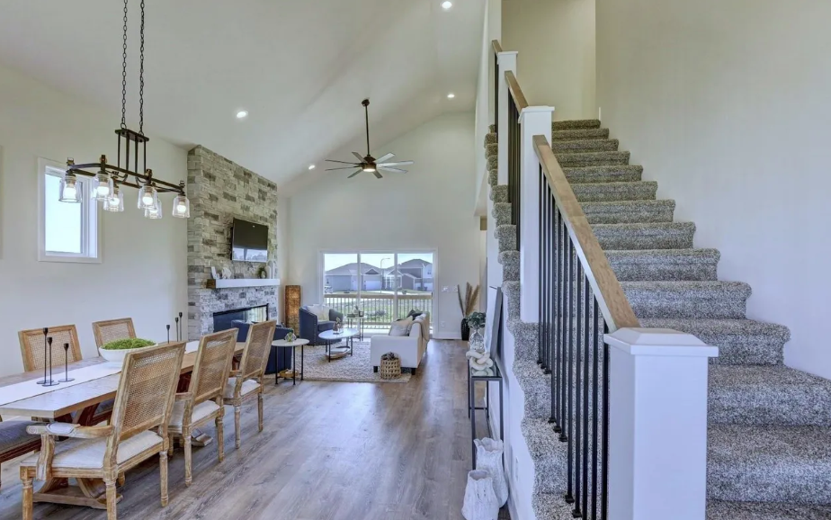 Open-concept living and dining area with vaulted ceiling, stone fireplace, ceiling fan, and staircase with carpeted steps and wooden handrail.