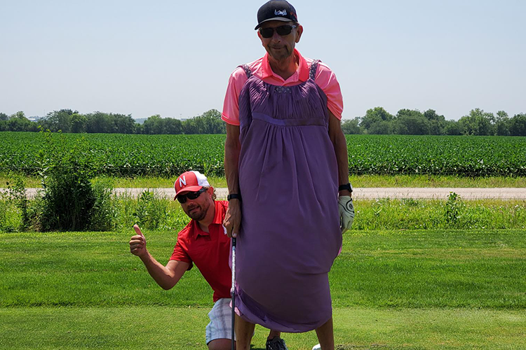 Two men, one standing with a purple golf umbrella and the other crouching beside him, on a golf course with greenery under a clear blue sky.