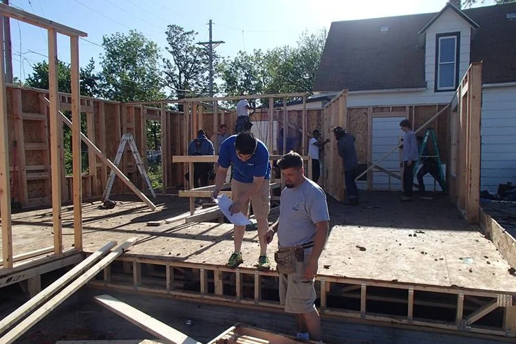 People working on building a wooden house or extension, with framing and construction materials visible.