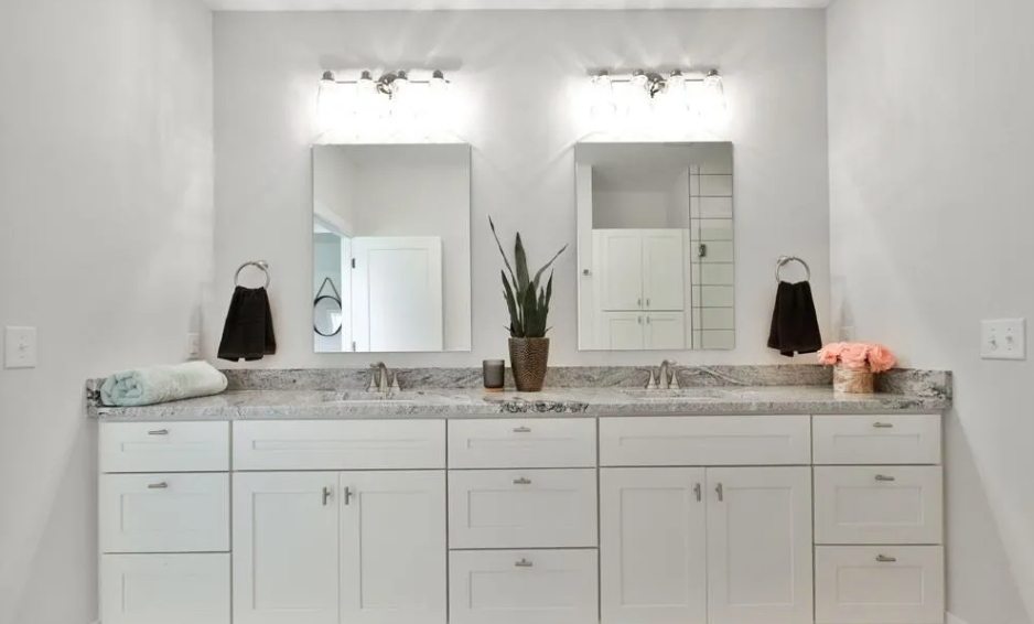 Bathroom vanity with two mirrors, a granite countertop, and two sinks. There are black towels hanging on each side, a potted plant in the center, and towels and flowers on the right side.