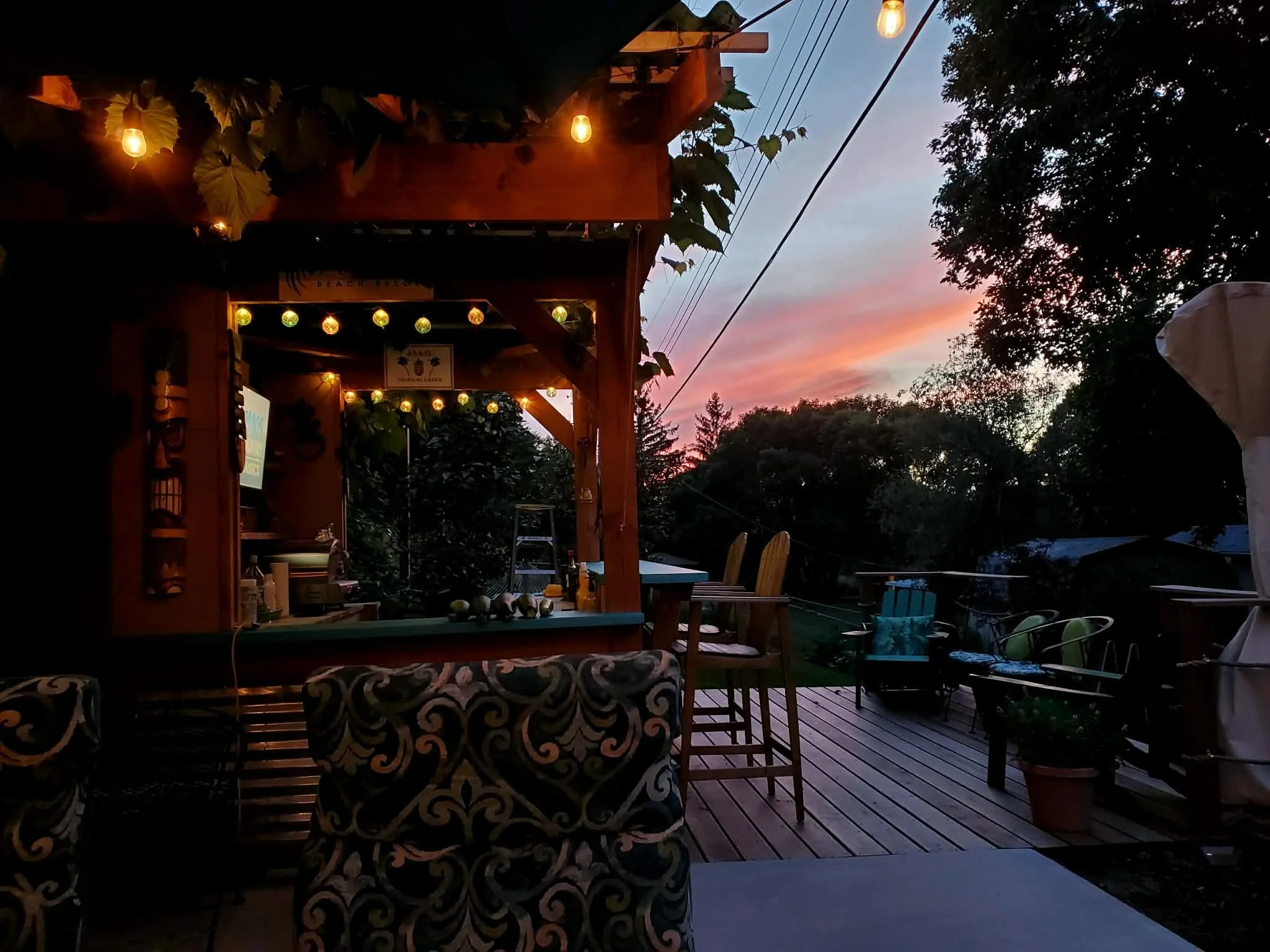 An outdoor deck area during sunset with string lights, wooden furniture including chairs and bar stools, potted plants, trees, and a colorful sky with pink and blue hues.