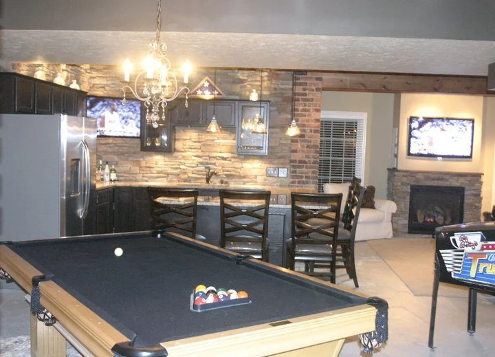 A recreational room featuring a pool table, kitchen area with bar stools, chandelier, and a living area with a fireplace and television.