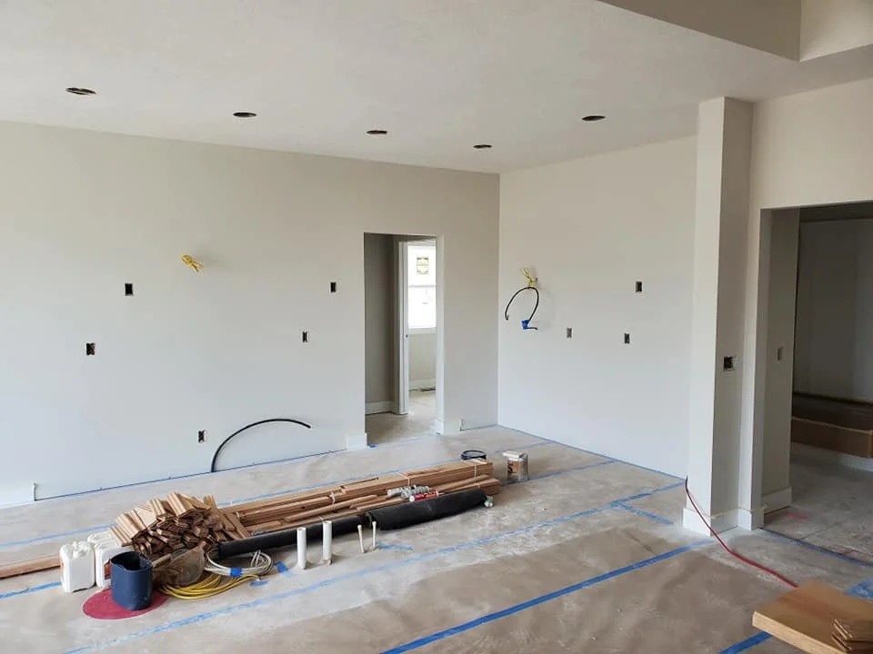 Interior of a house under construction with white painted walls, recessed ceiling lights, electrical wiring, and construction materials on the floor.
