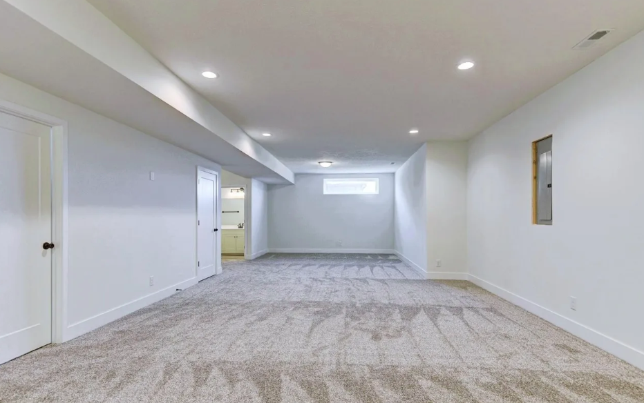 Empty basement room with white walls, beige carpet, and recessed ceiling lights, featuring a small horizontal window and an open closet with shelves.