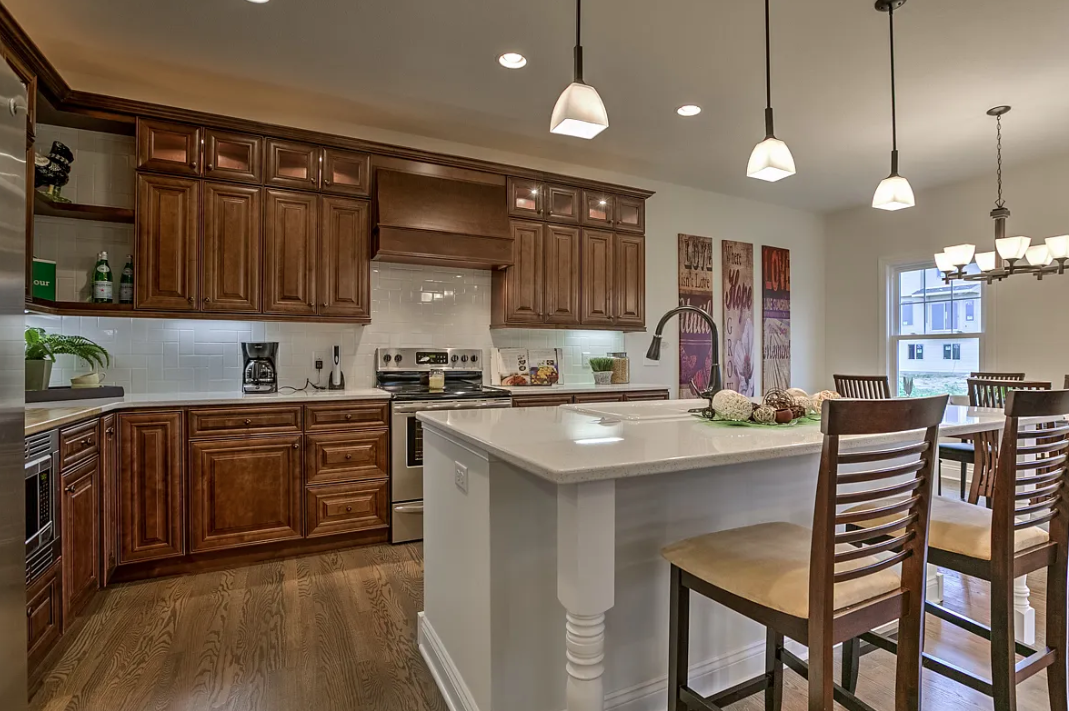 Kitchen with wooden cabinets, white countertops, white backsplash, stainless steel appliances, island with bar stools, and dining area with a chandelier and window.