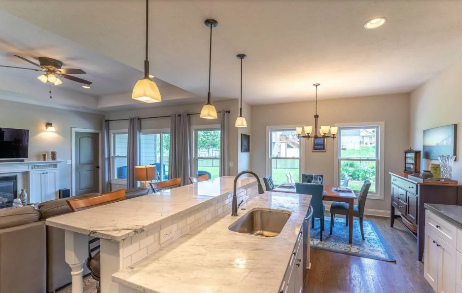 Open-concept living and dining room with a kitchen island, pendant lights, a dining table with chairs, a sideboard, large windows, and various light fixtures.