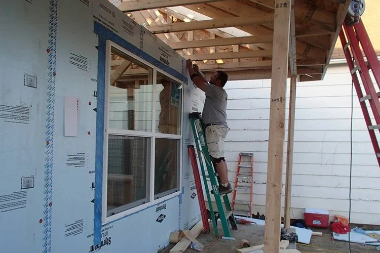 A person standing on a ladder installing or working on the roof of a house under construction, with building materials and tools around.