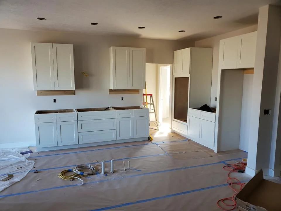 Kitchen under construction with white cabinets and no appliances, covered floor, and construction materials and wires on the floor.