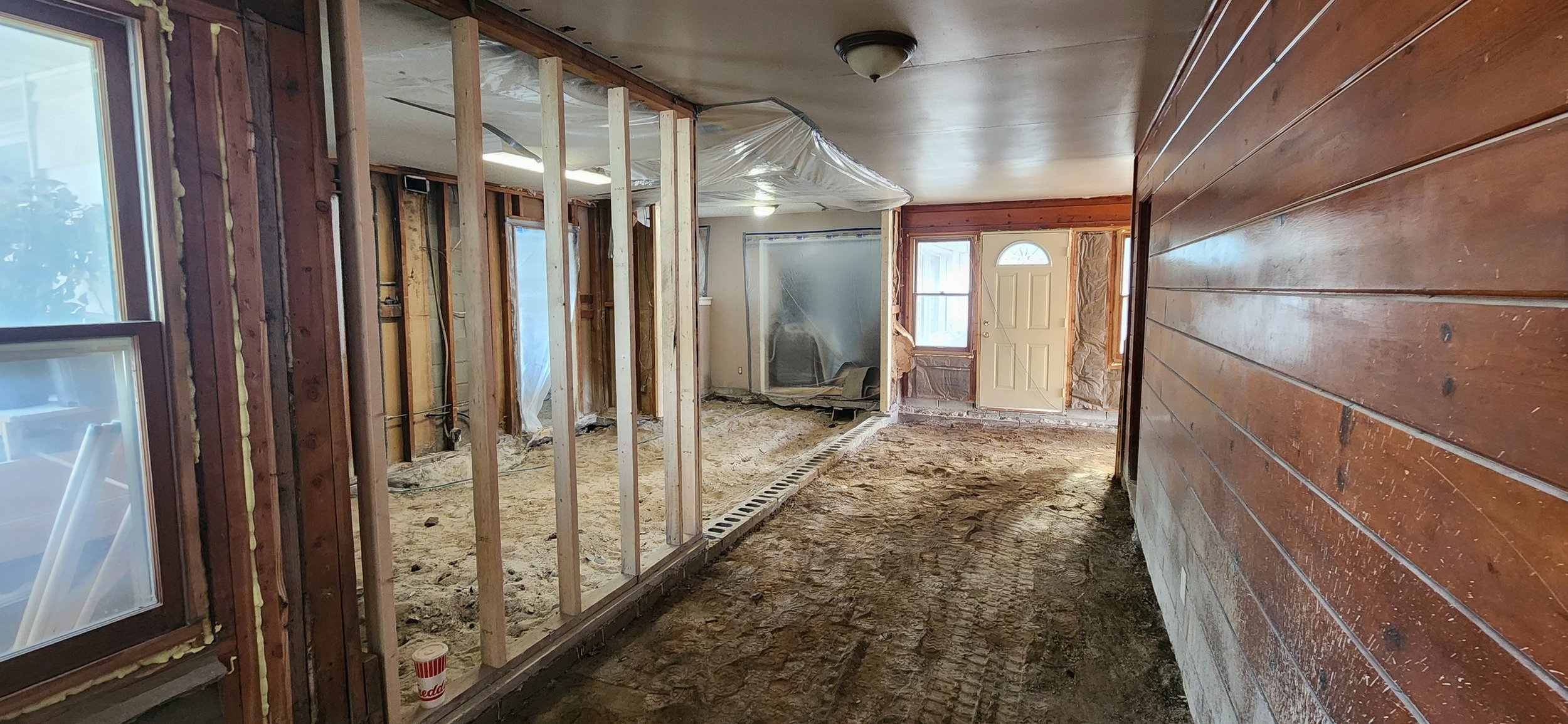 Interior of a house undergoing renovation, with exposed wooden framing, dirt floor, and partially installed wall and window panels.