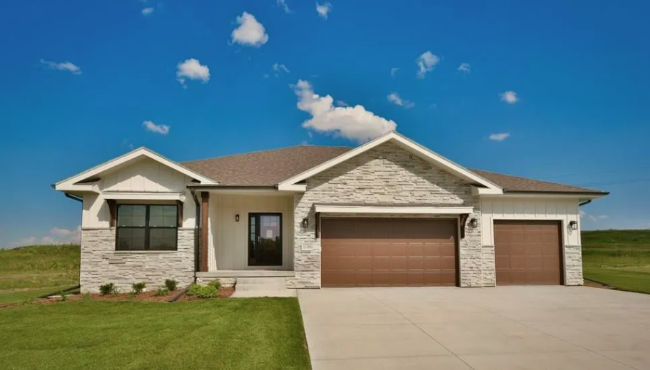 A modern single-story house with a stone and white siding exterior, a brown garage door, and a front porch, set against a blue sky with scattered clouds.