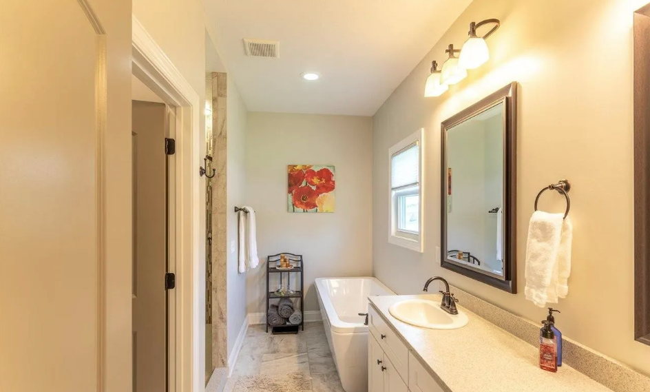 Bathroom with a white vanity, a mirror, a towel rack, a bathtub, a small window, and a small cart with folded towels and toiletries.
