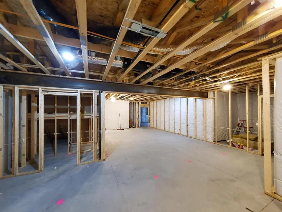 Interior view of a house under construction, showing wooden framing, exposed ceiling joists, and electrical wiring, with concrete flooring and some construction tools and equipment.
