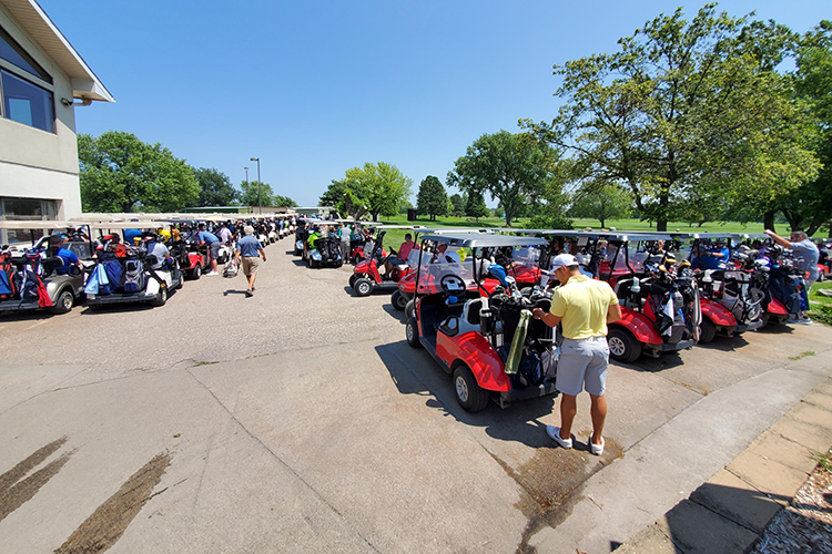 A large group of people at a golf course parking lot, preparing to play golf with numerous golf carts lined up.