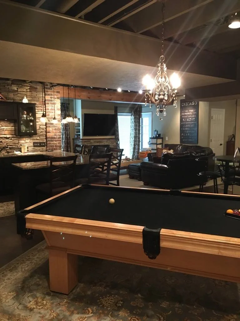 Living room with pool table, leather couch, chandelier, brick accent wall, flat-screen TV, and bar area.