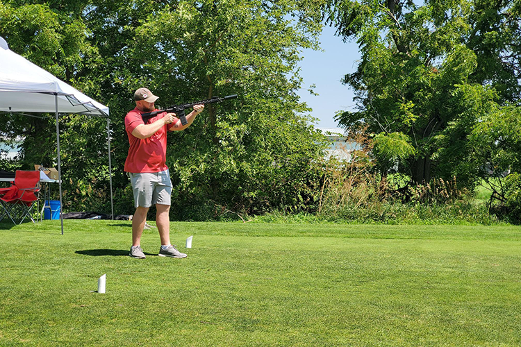A man in a red shirt and beige shorts is aiming a rifle at a shooting range on a grassy field, with trees and a blue sky in the background, and a white canopy tent to the left.