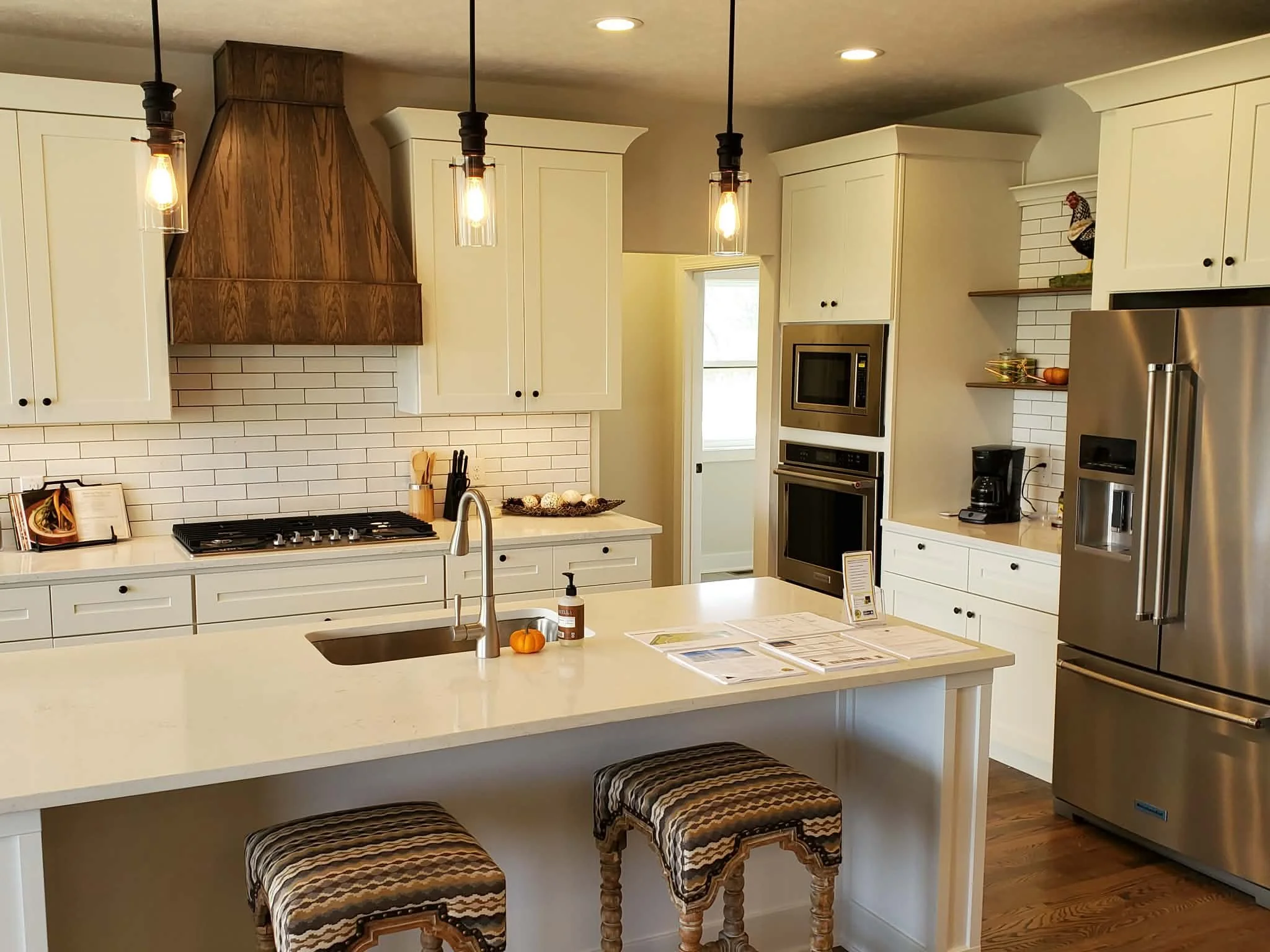 Modern kitchen with white cabinets, stainless steel refrigerator, built-in microwave and oven, island with two patterned stools, pendant lights, white subway tile backsplash, and a window. Small decorative pumpkin and bottles on the island.