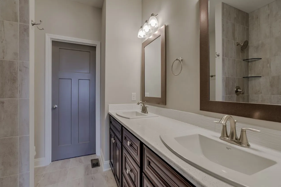 Bathroom with dual sinks, large mirror with brown frame, wall-mounted light fixture, gray door, beige tiled floor, and shower with beige tile walls and glass shelves.