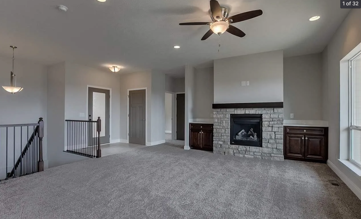 Empty living room with a carpeted floor, a ceiling fan, a fireplace with a stone surround, built-in cabinets, and large windows.