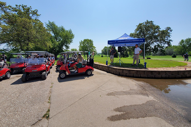 Golf carts parked near a stage with a speaker and an audience at a golfing event on a sunny day.