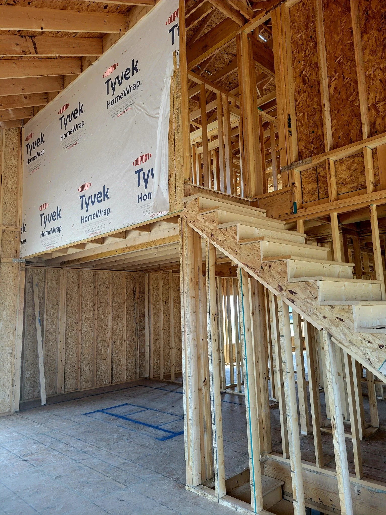 Interior view of a house under construction, showing wooden framing, stairs, and a large sheet of insulation material labeled "Tyvek HomeWrap."