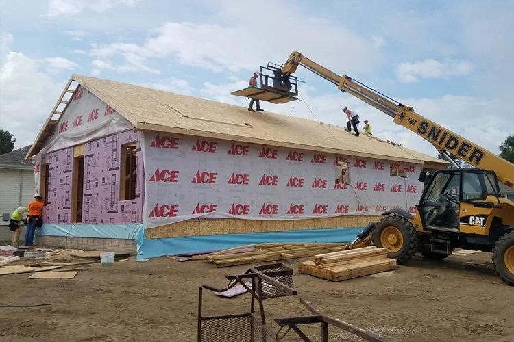 Construction workers working on a house under construction, with a brick exterior wrapped in pink house wrap, a crane lifting a worker on a platform to the roof, and building materials on the ground.