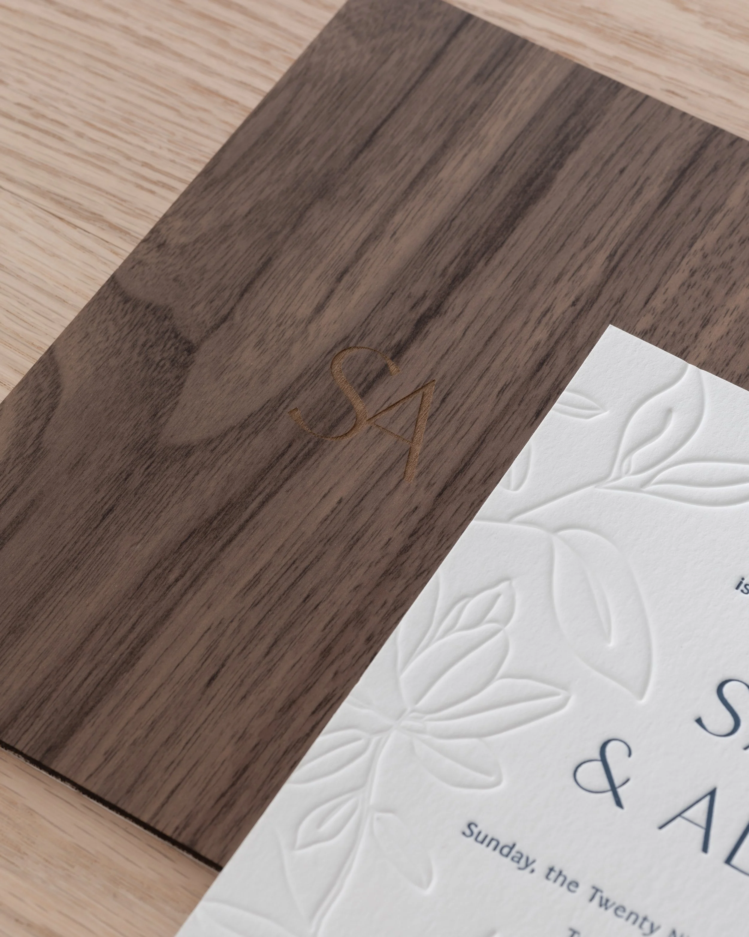 A close-up of a wooden menu or card with the initials 'SA' engraved on it, partially covering an embossed floral design on a white paper.