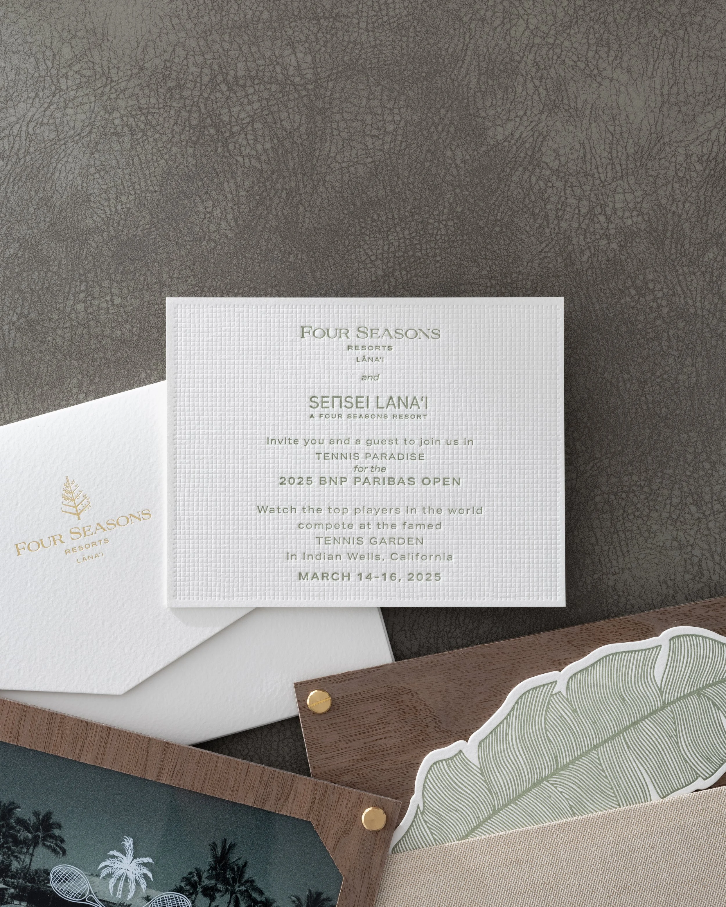 Invitation card for the 2025 BNP Paribas Open tennis tournament at Indian Wells, California, from March 14 to 16, 2025, placed on a textured gray surface, with other items including a white envelope, a wooden tray with pictures and a large leaf illus