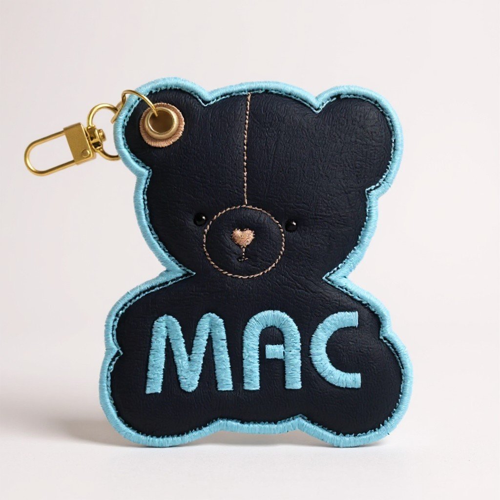 Black bear-shaped keychain with blue outline and embroidered "MAC" on chest, attached to a gold clip.