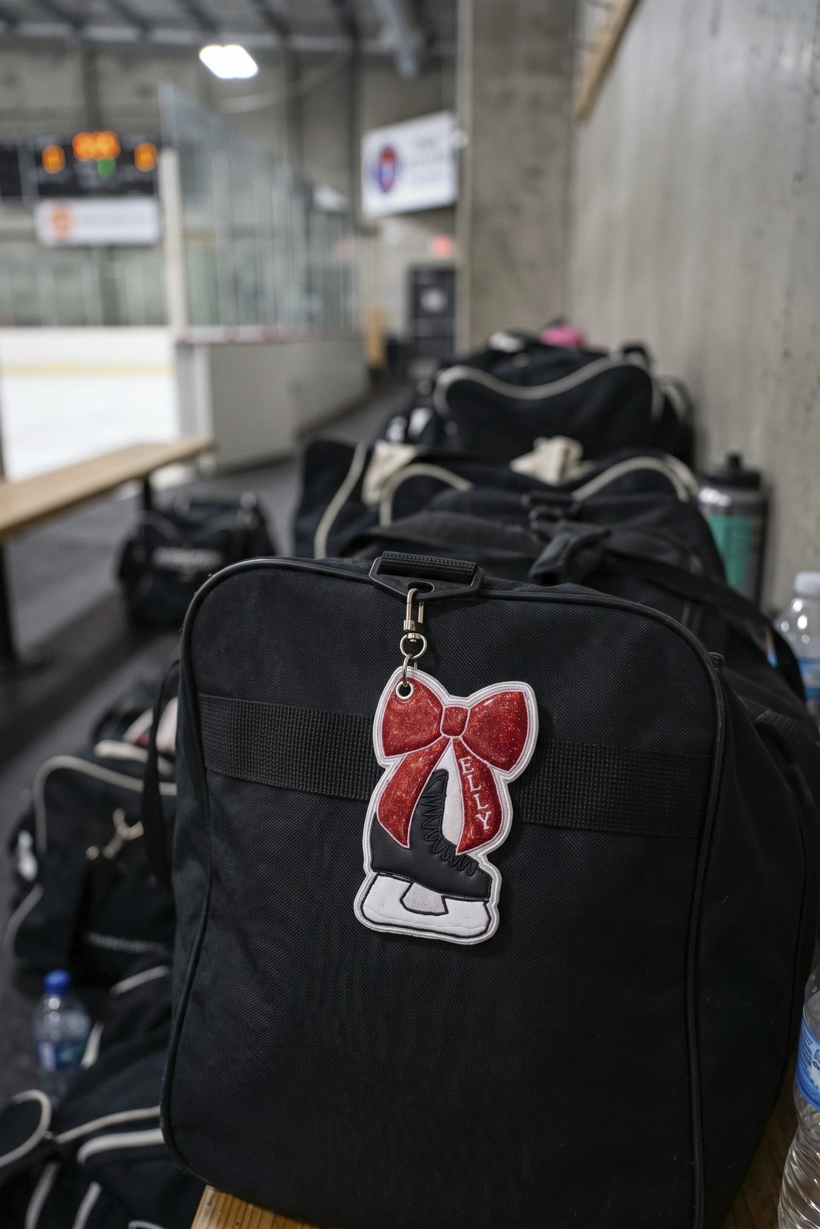 Image of black hockey bags all together in ice rink with one bag clearly standing out with embroidered hockey skate bow bag charm hanging from zipper.