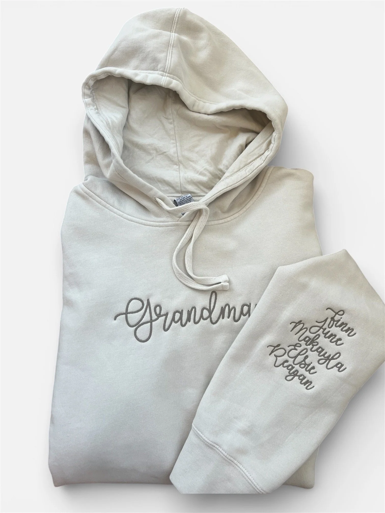 White hoodie sweatshirt featuring personalized warm brown script embroidery. The hoodie says 'Grandma.' The sweatshirt sleeve shows the names of grandkids.