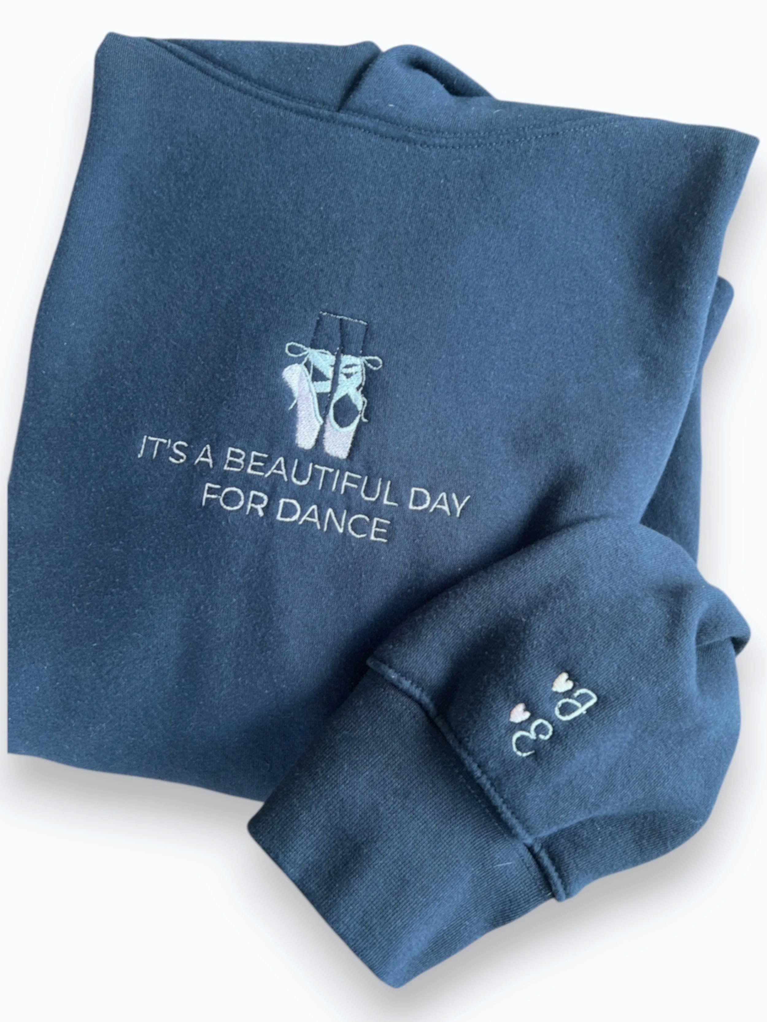 A vintage black sweatshirt with embroidered ballet slippers and the phrase "It's a beautiful day for dance" on the front, folded with the sleeve showing on the side with embroidered heart and initials.