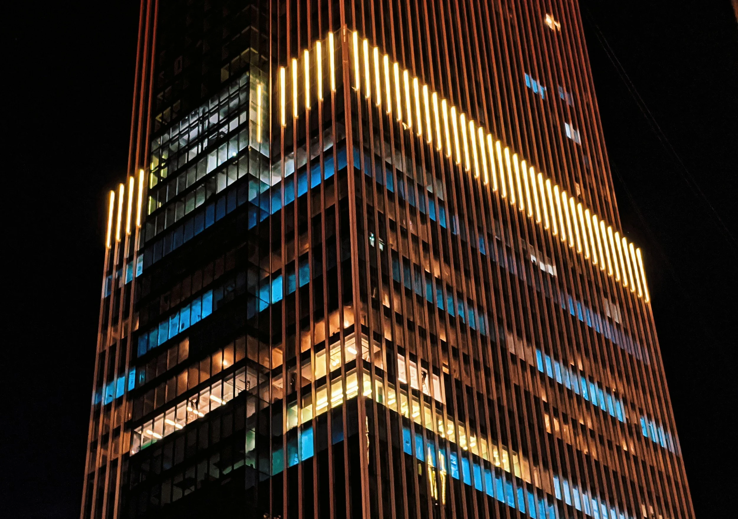Nighttime photo of a modern skyscraper with vertical wooden slats, illuminated by yellow and blue lights.