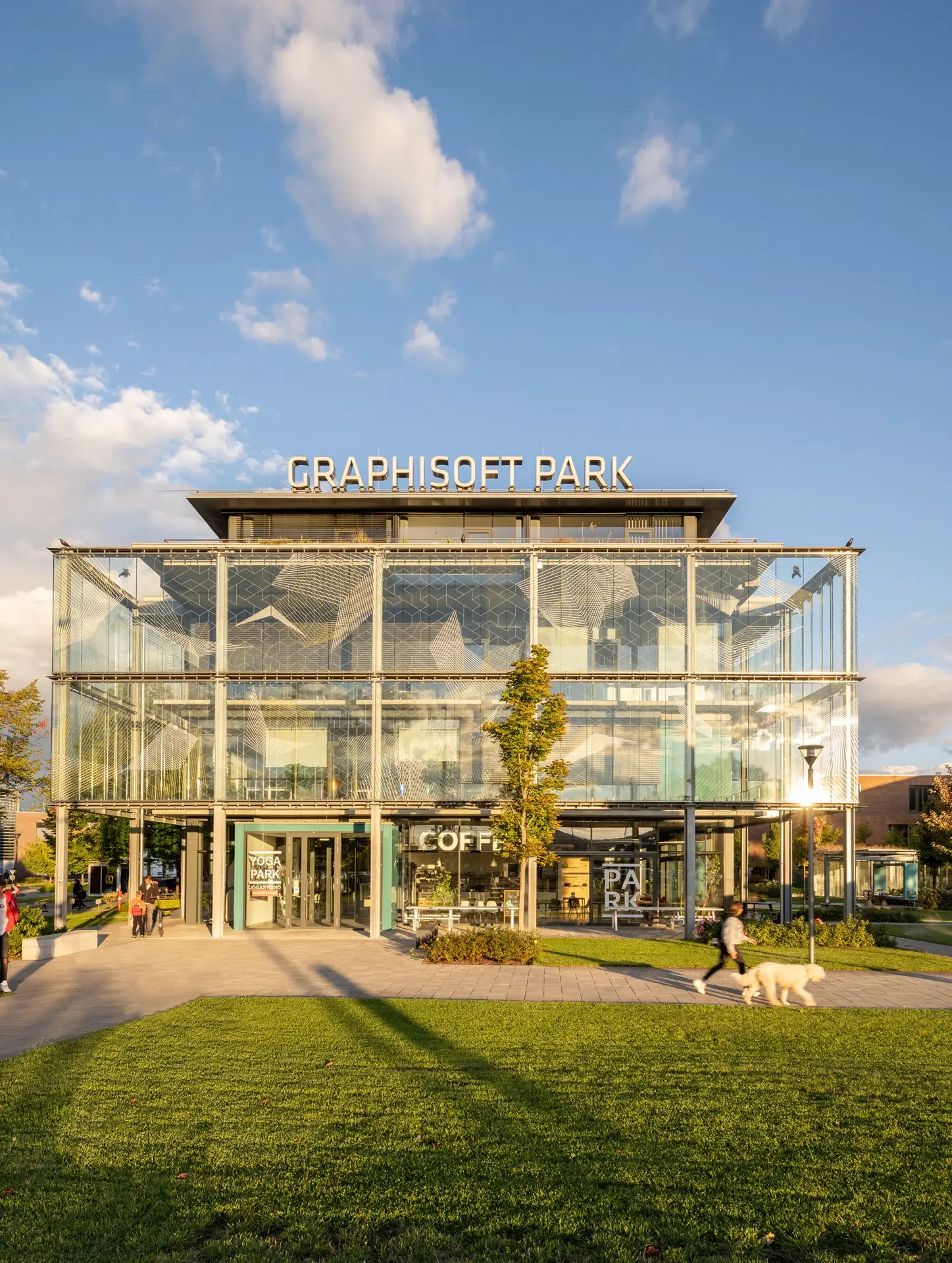 A modern glass building with the sign 'GRAPHISOFT PARK' on top, with people walking outside, some with dogs, and a grassy area in the foreground, under a partly cloudy sky.