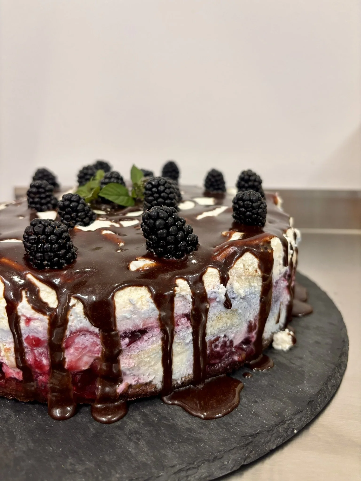 Chocolate drizzled berry cake topped with blackberries and mint leaves on a black slate serving platter.