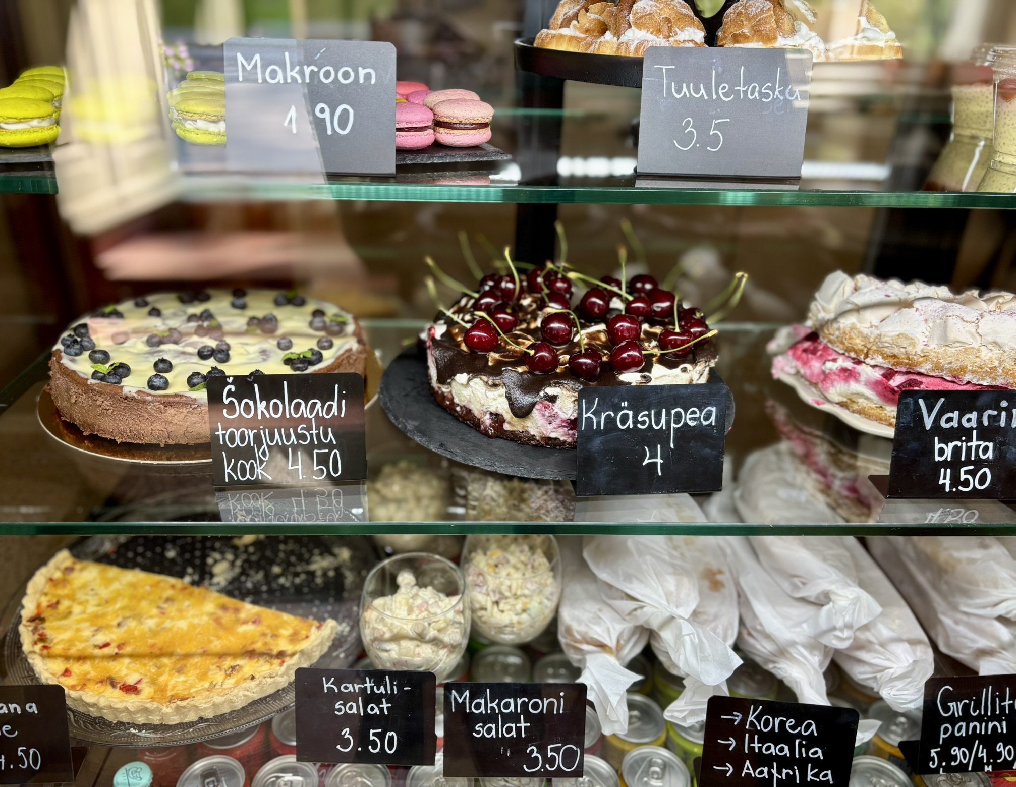 Display case with various desserts and baked goods including chocolate cake with blueberries, cherry cake with cherries, fruit pie, macarons, salads, and wrapped sandwiches in a bakery or pastry shop.