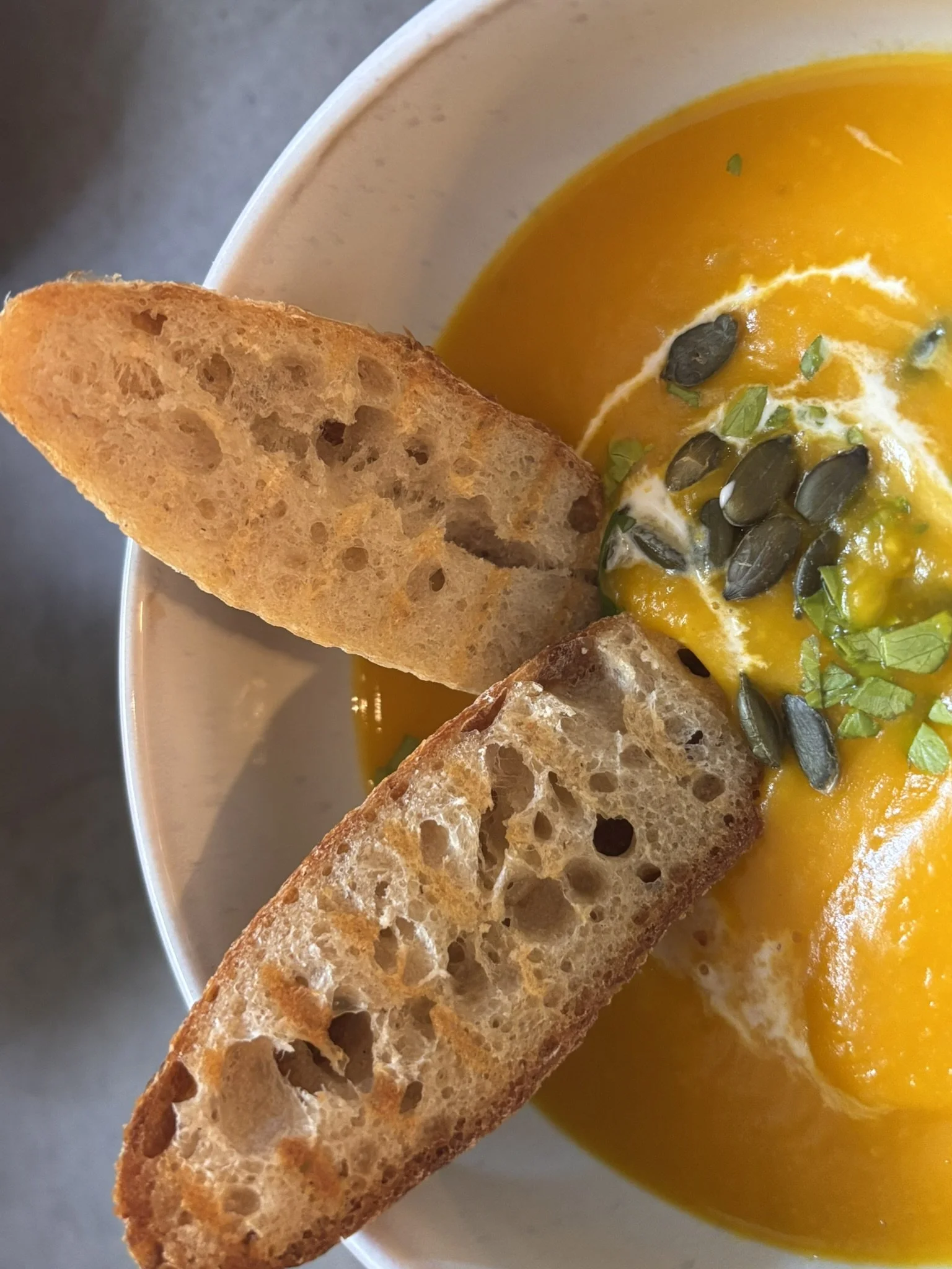 Close-up of a bowl of bright orange soup topped with pumpkin seeds, chopped greens, and a swirl of cream, accompanied by two slices of toasted bread.