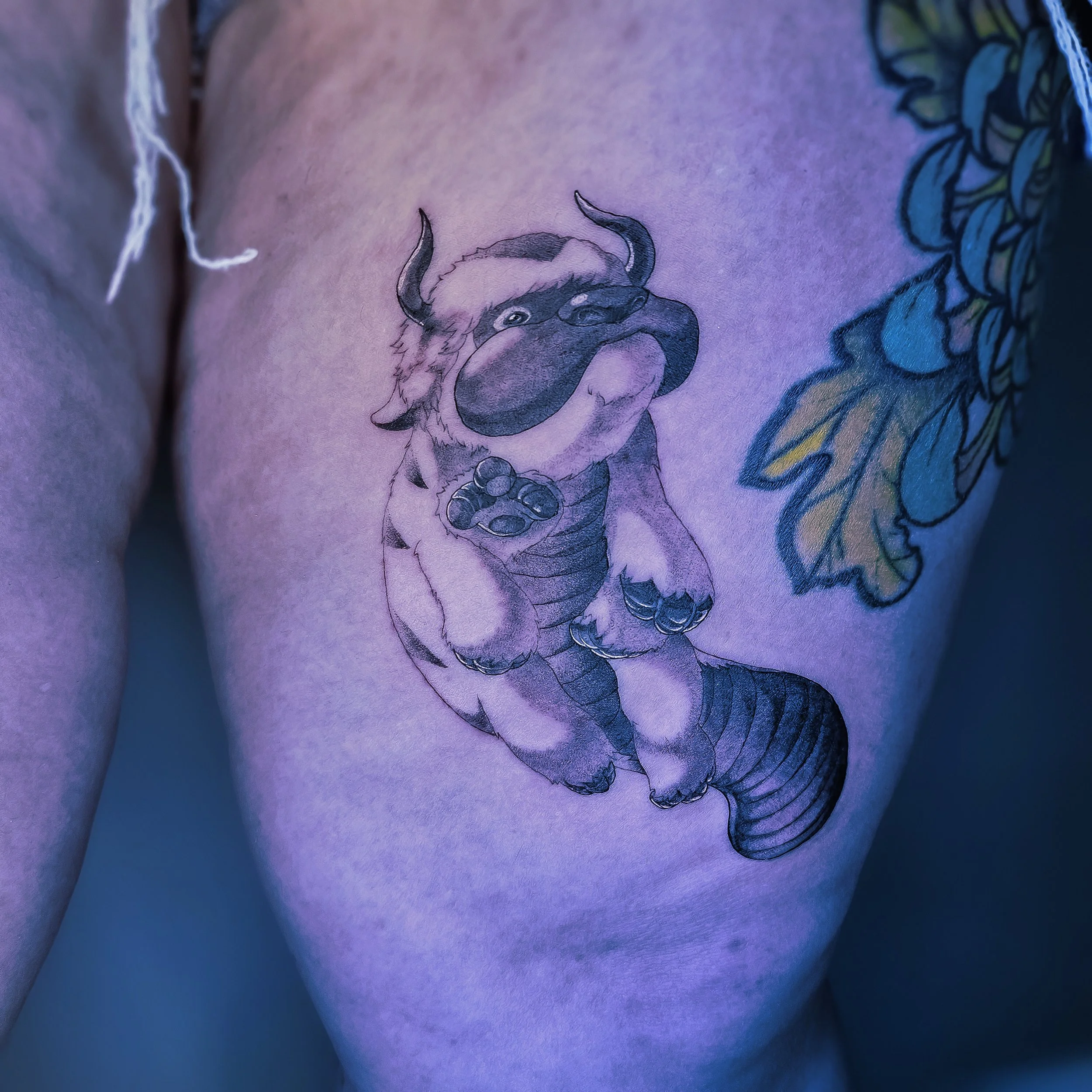 A tattoo of a cartoon-style creature with the body of a tiger, the face of a dog, horns, and a wagging tail on a person's chest, alongside colorful leaves.