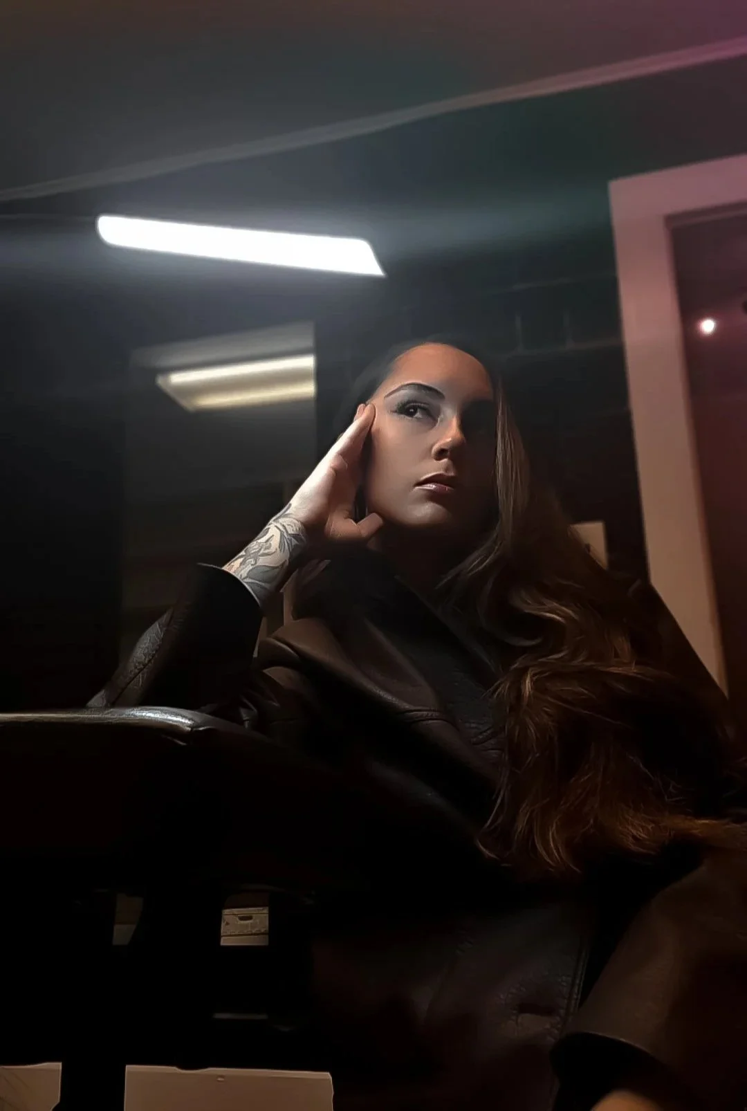 A woman with long brown hair and tattooed arm sitting in a dark room, resting her head on her hand, with reflective lighting overhead.