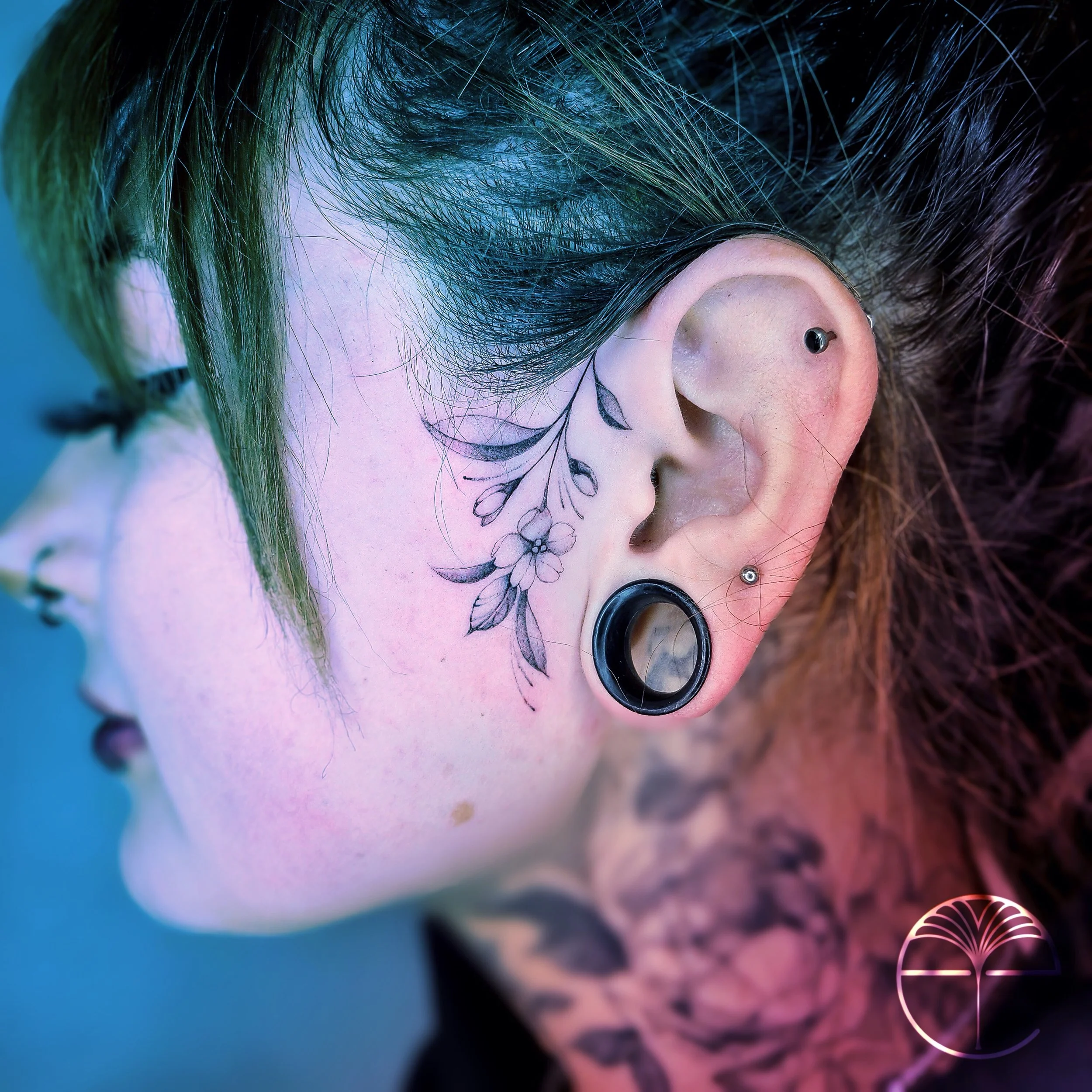 Close-up of a person's ear with multiple piercings, including a black gauged earlobe, a snuggie piercing, and a helix piercing, with floral tattoos on the skin next to the ear. The person has short, multi-colored hair and a neck tattoo visible in the