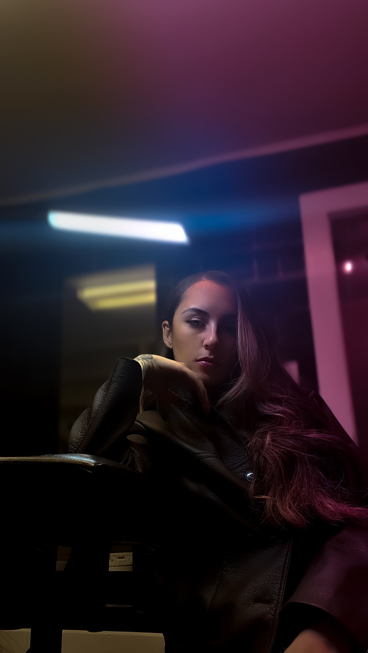 A young woman with long wavy hair and makeup sits indoors under colorful neon lighting in a dimly lit room.