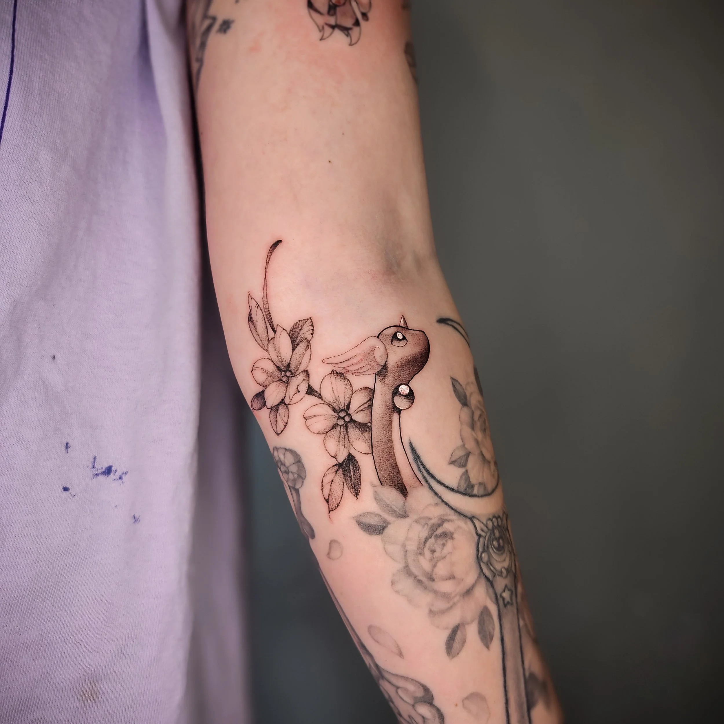 Close-up of a person's arm with a black and gray tattoo of a bird with flowers, including cherry blossoms, on the forearm.
