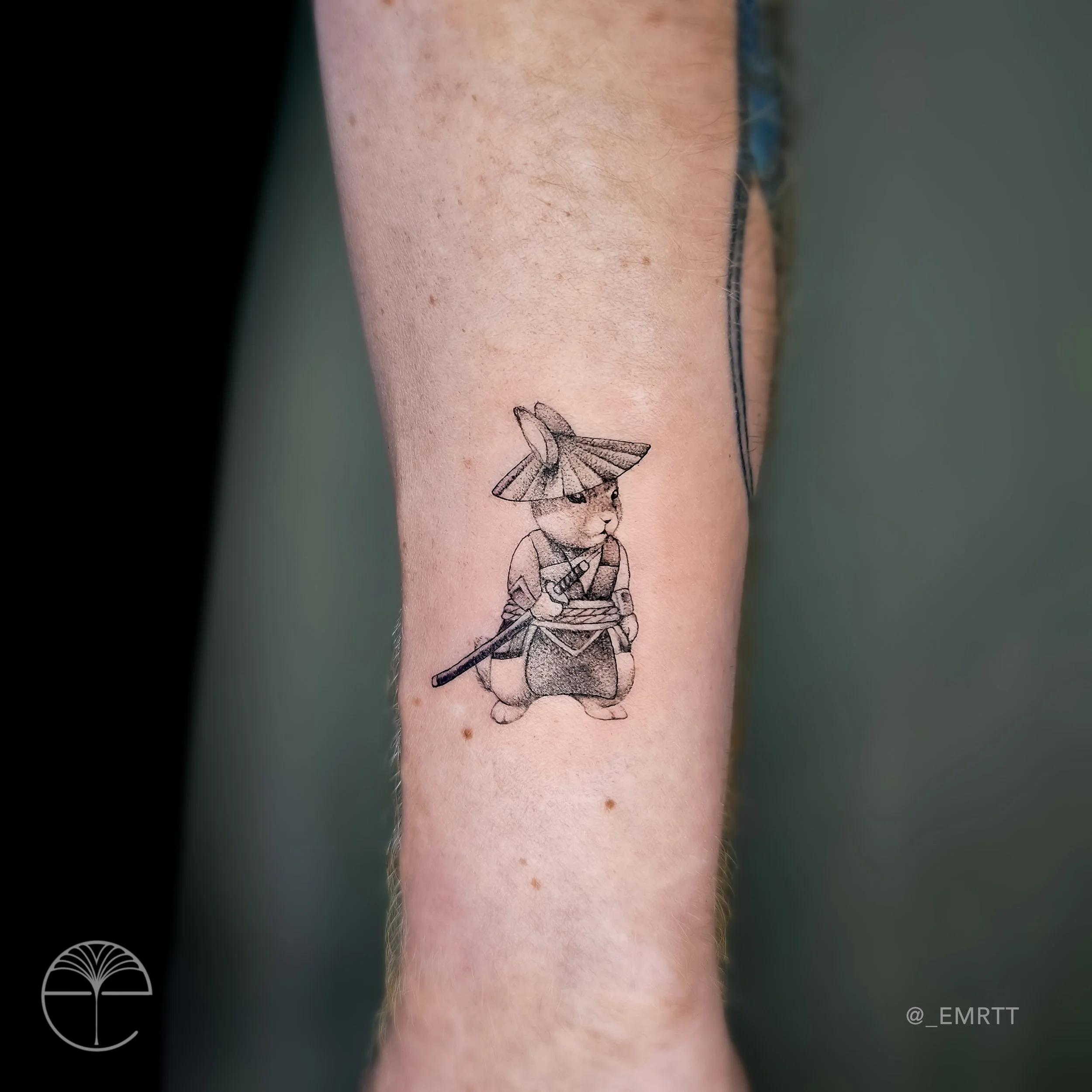 Tattoo of a cute anthropomorphic bear dressed as a samurai, holding a sword and wearing a traditional straw hat on a person's forearm.