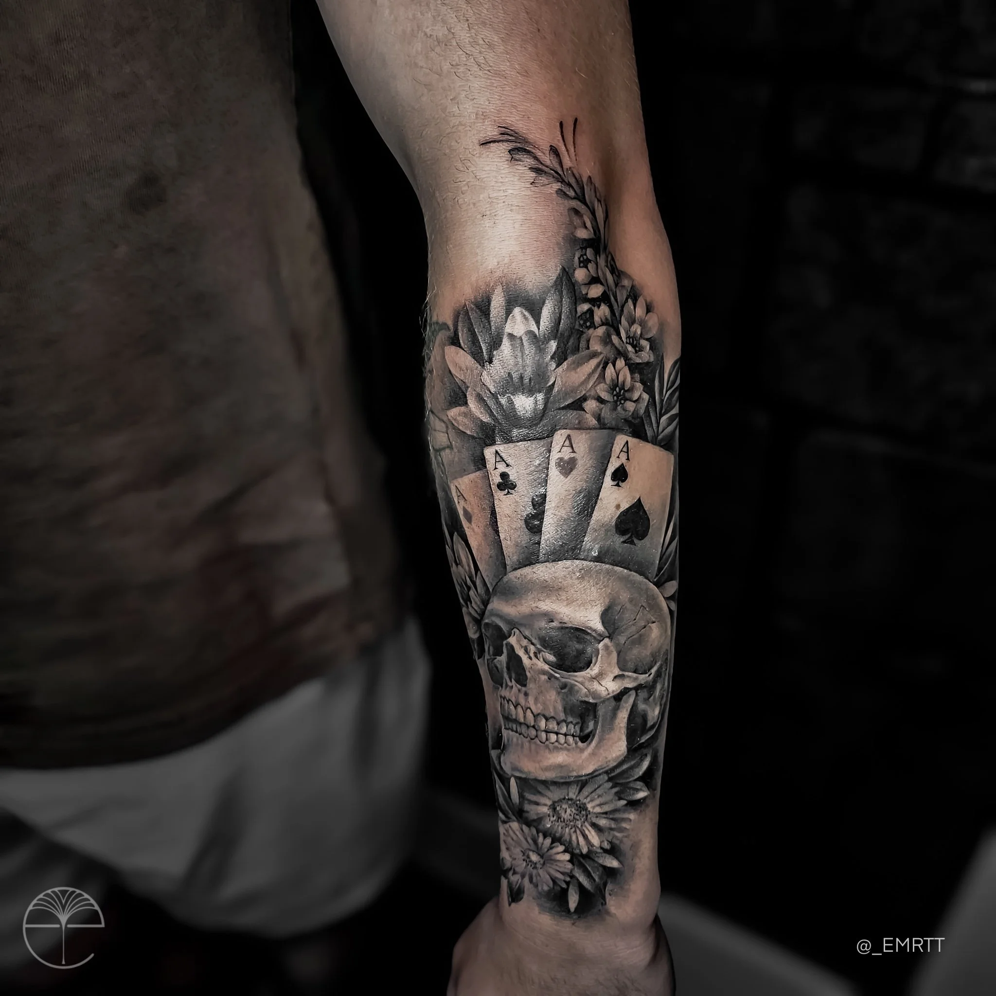 Black and gray tattoo on a forearm featuring a skull, playing cards showing aces, flowers, and a bee.