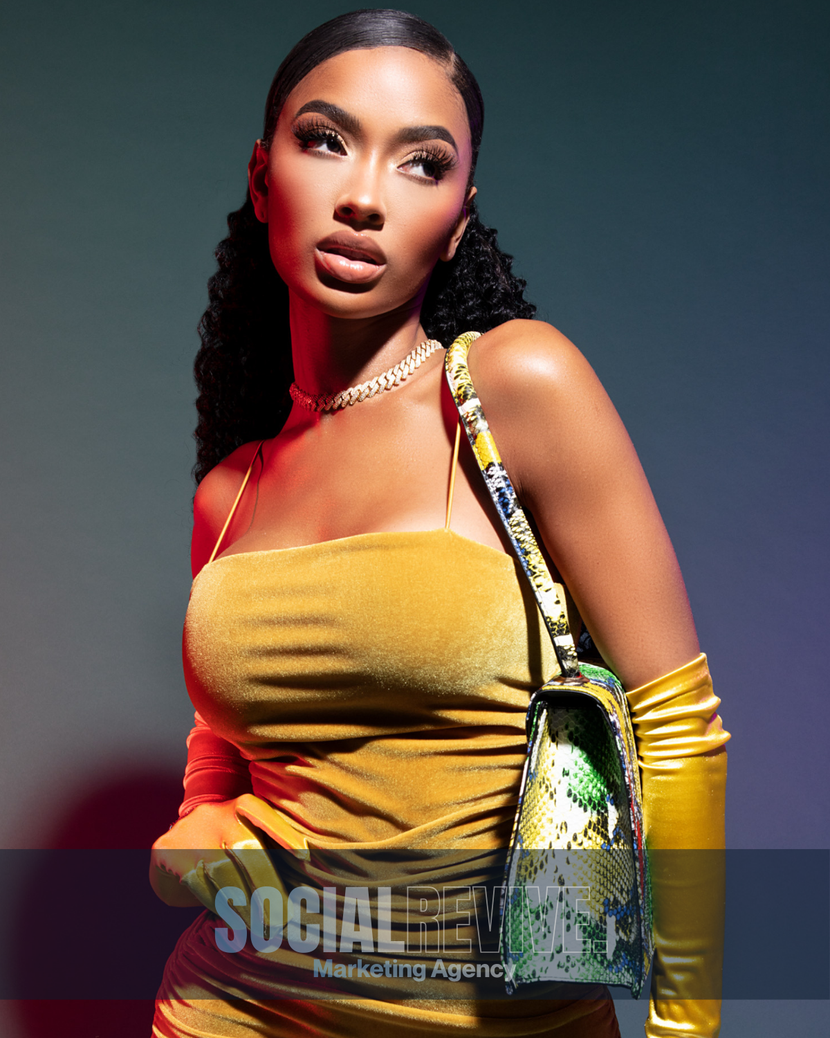 A woman with dark hair styled in waves, wearing a yellow dress, gold gloves, a gold chain necklace, and holding a colorful patterned handbag, poses against a dark background.