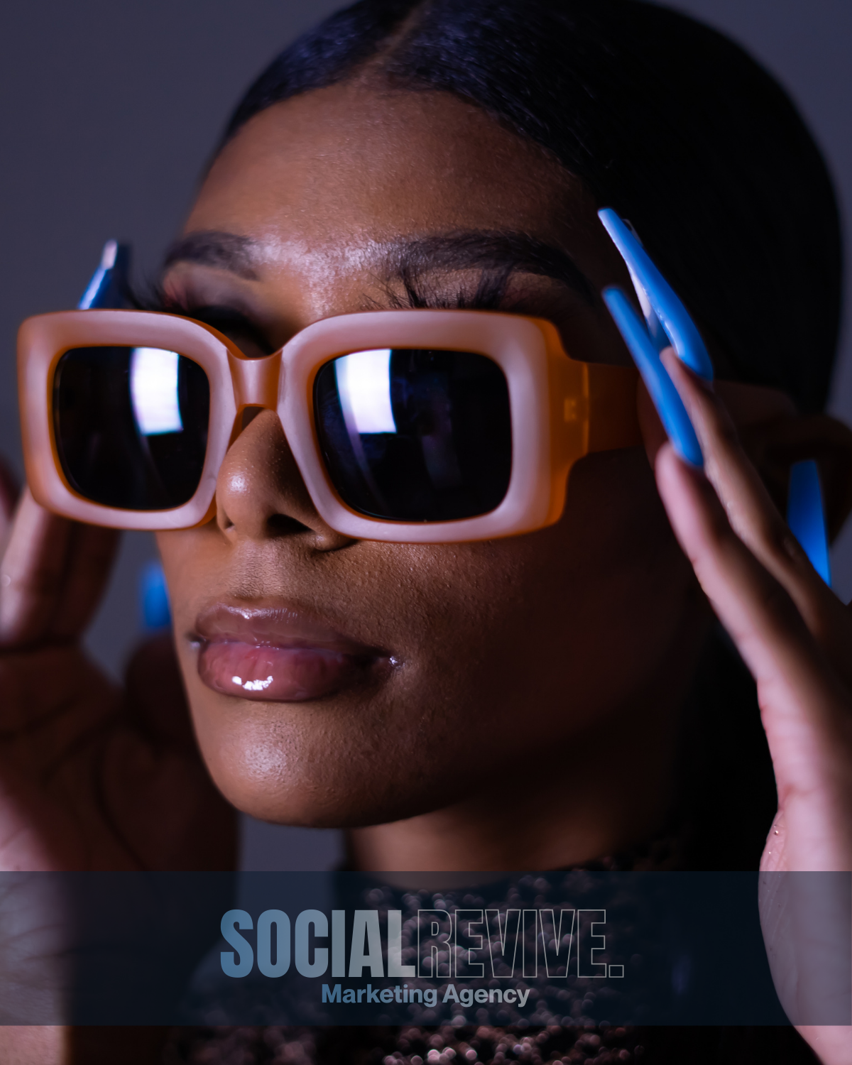 Close-up of a woman wearing large orange sunglasses with reflective lenses, adjusting them with her hands. She has dark hair and is wearing glossy lipstick. The background is blurred, and there is a logo for Social Revive Marketing Agency at the bottom.
