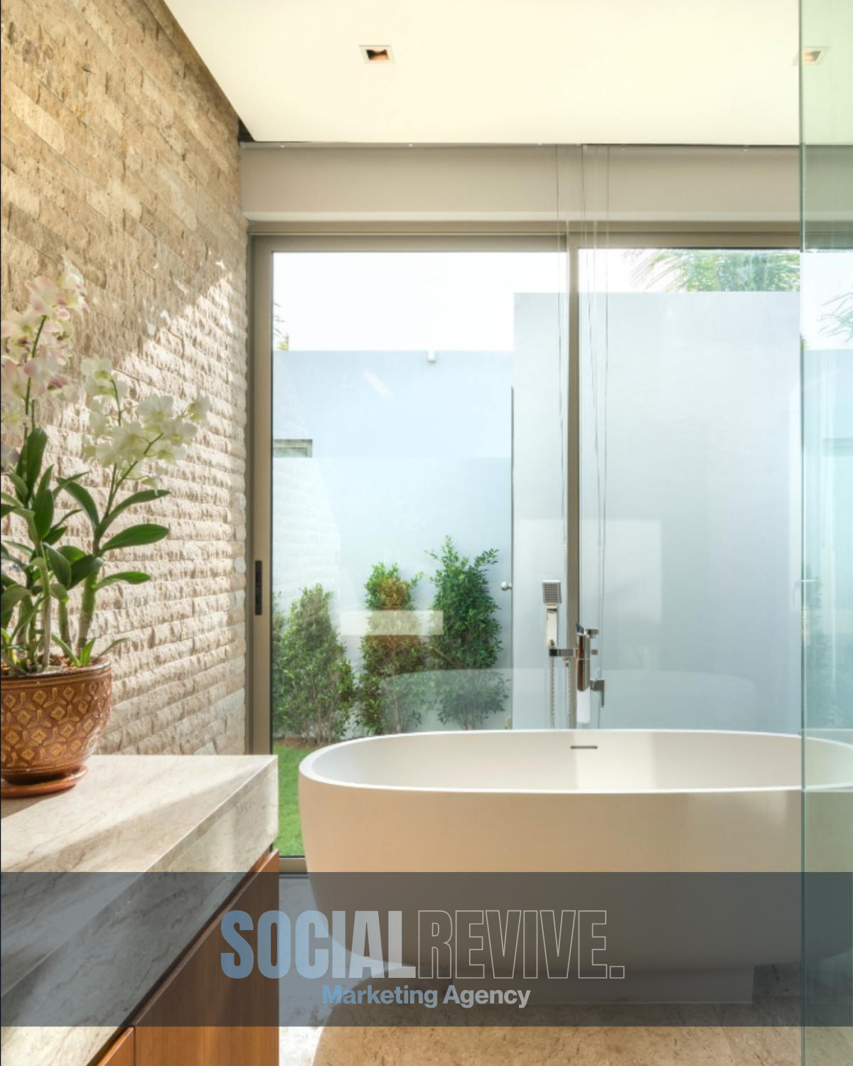 Modern bathroom with a freestanding bathtub, large glass window showing outdoor plants, exposed brick wall, and a potted orchid on a marble countertop.