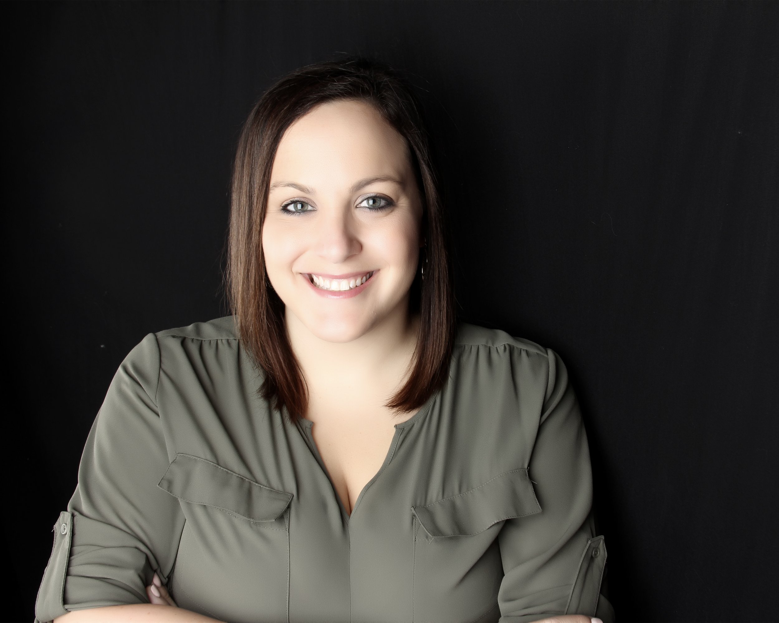 A woman with shoulder-length brown hair, smiling, wearing a gray shirt, standing against a black background.