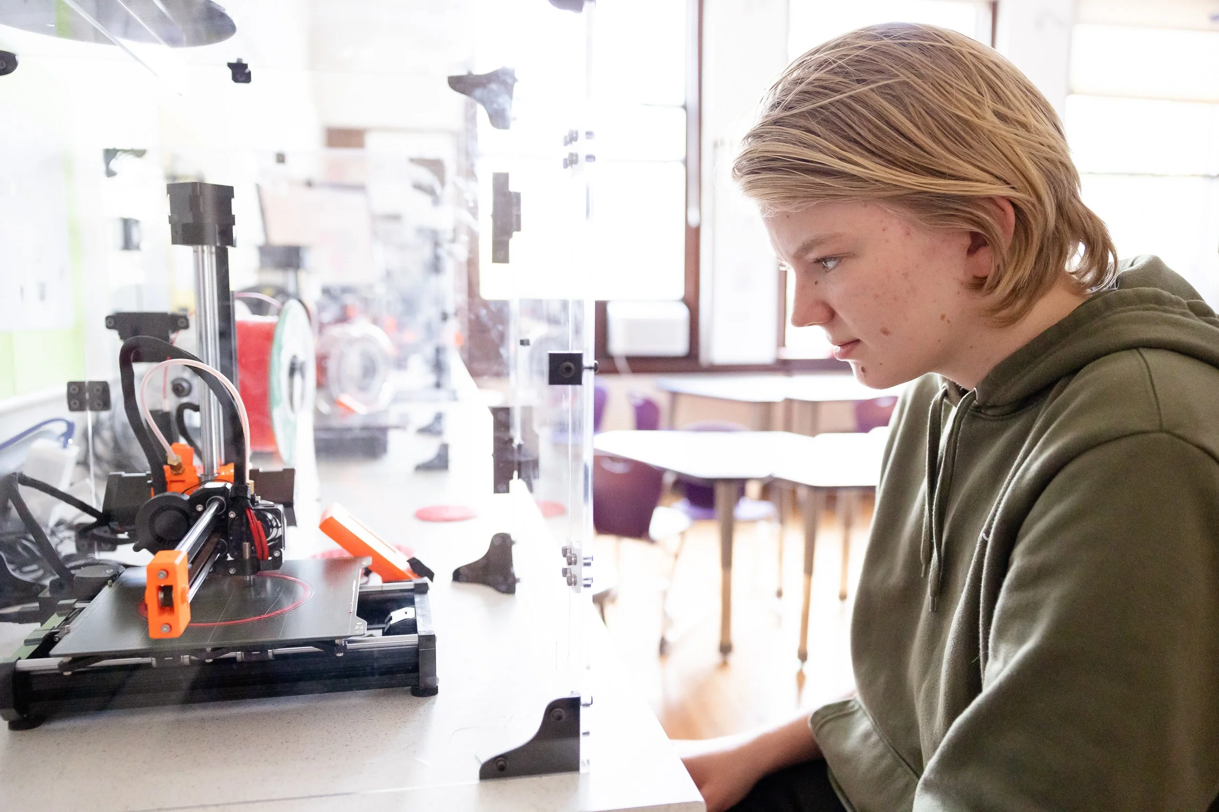 A middle school student sitting at a table and working on a 3D printer at Wellan Montessori school.